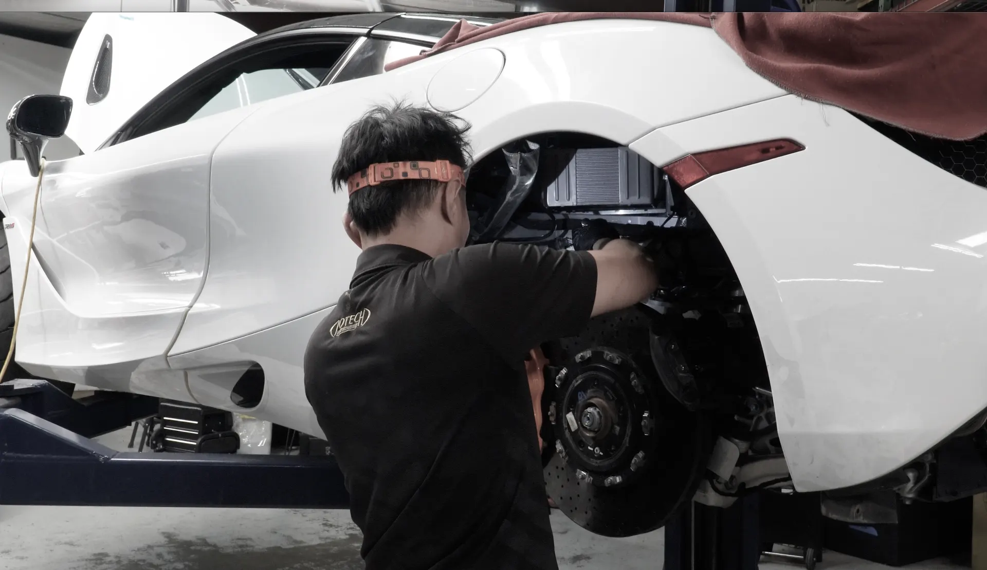A mechanic works on the rear brake and suspension of a white McLaren supercar on a lift in a performance shop.